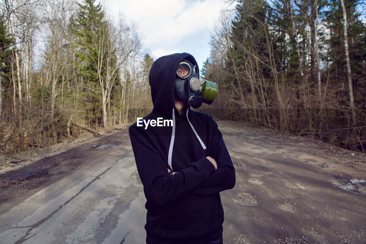 Guy stands in a post apocalyptic forest during the day wearing a gas mask during a coronavirus