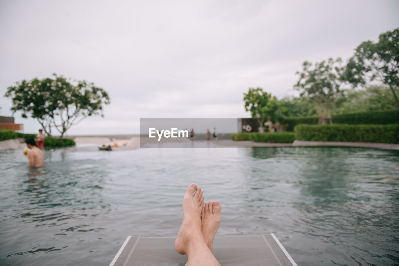 Low section of person floating on water in swimming pool with people in background against sky