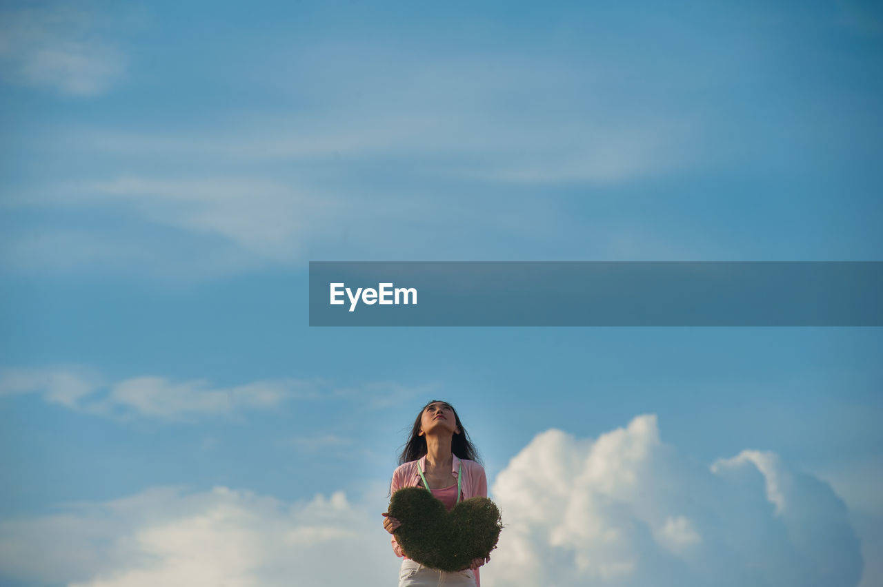 Woman holding heart shape while looking up against sky