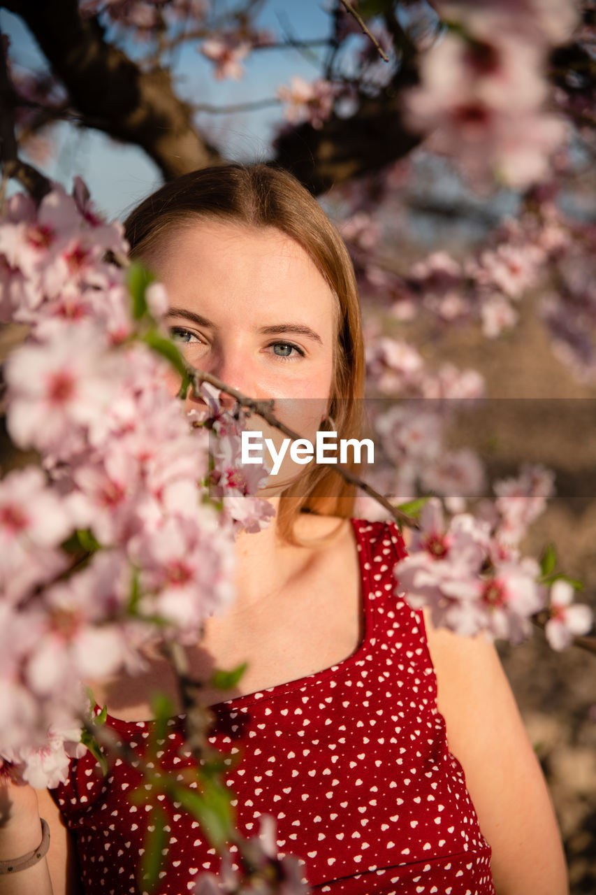 Blond woman with long hair posing under a flowering almond tree and looking at camera