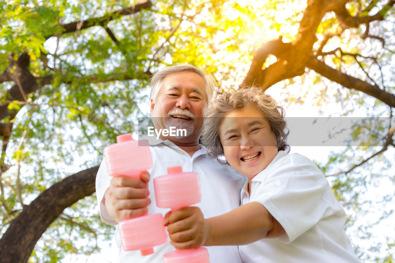 Portrait of smiling senior couple holding bottles against trees