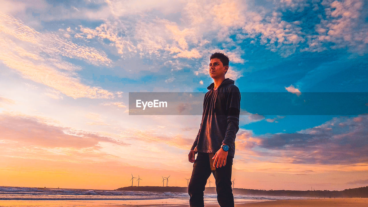 Young man standing at beach against sky during sunset