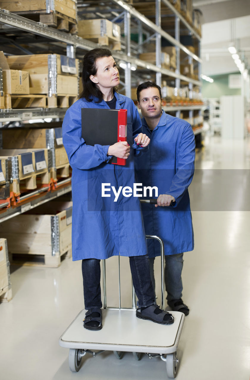 Male electrician pushing trolley while woman standing on it while working in factory