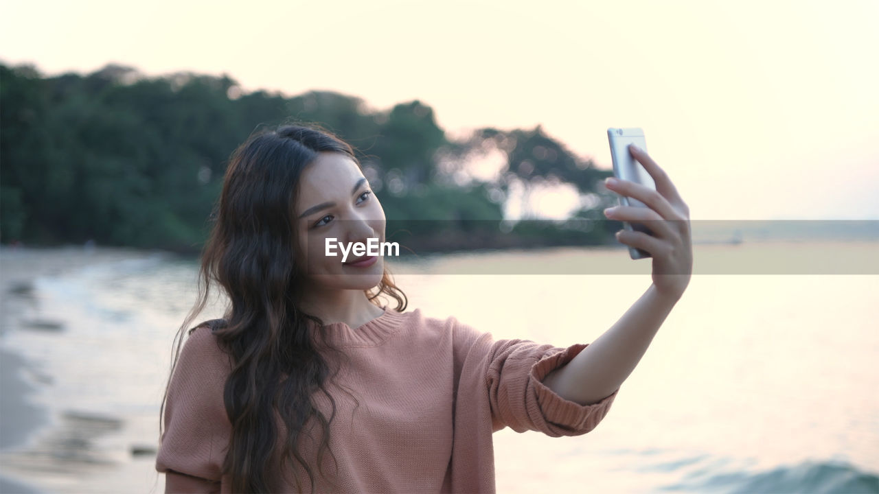 PORTRAIT OF BEAUTIFUL YOUNG WOMAN USING MOBILE PHONE AGAINST SKY