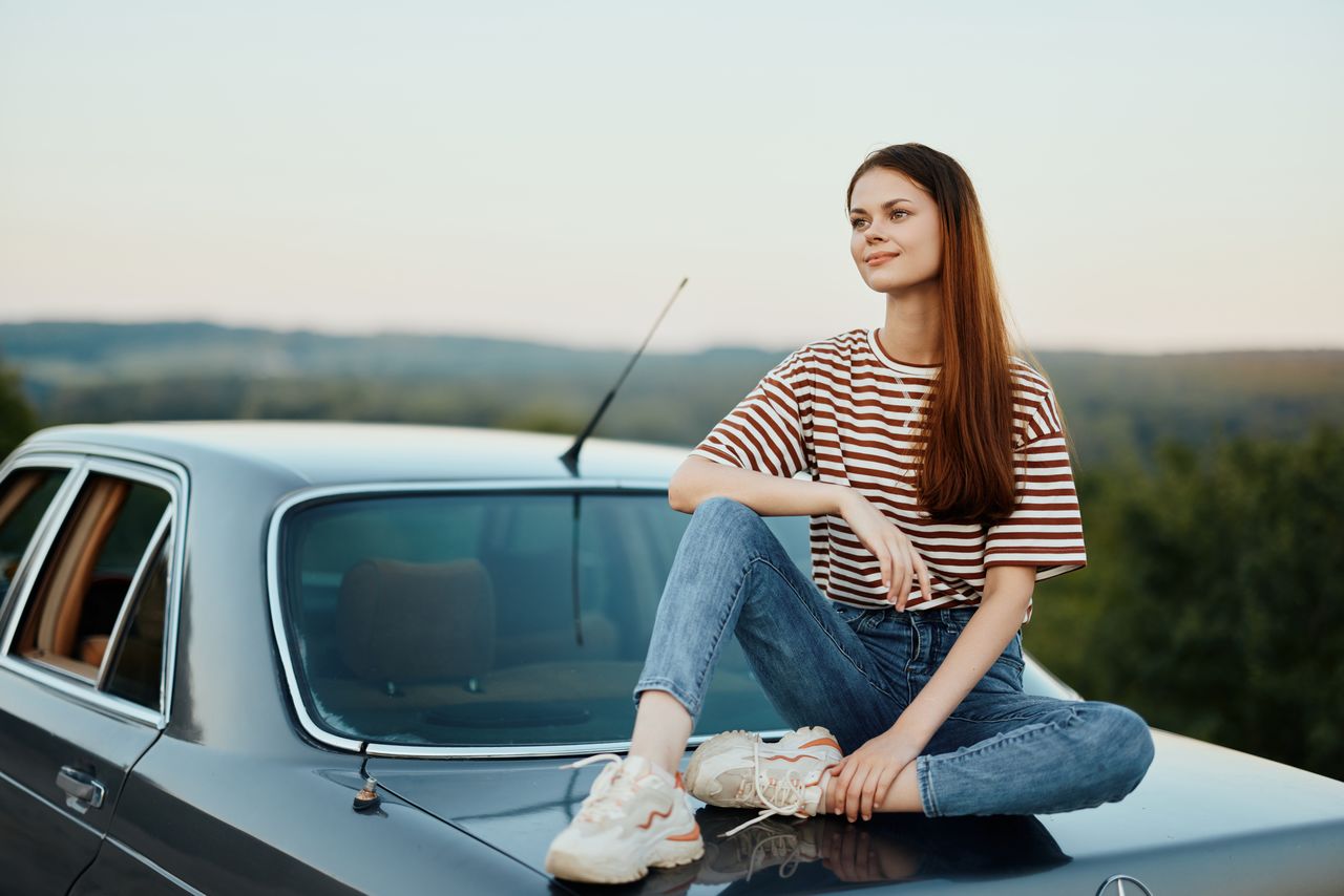 portrait of young woman sitting on car against sky