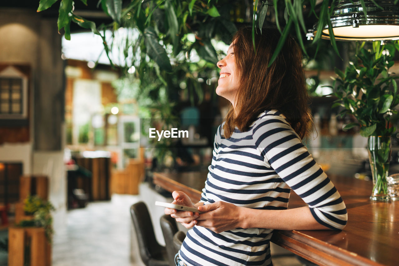 Young woman wearing striped longsleeve with mobile phone in hands in green eco cafe, city life