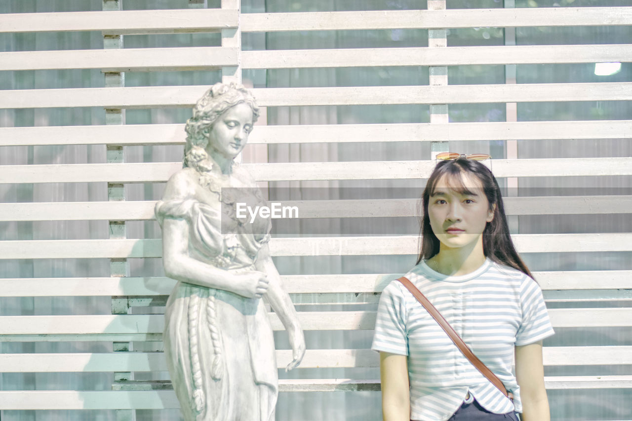 Portrait of young woman standing against sculpture by window outdoors