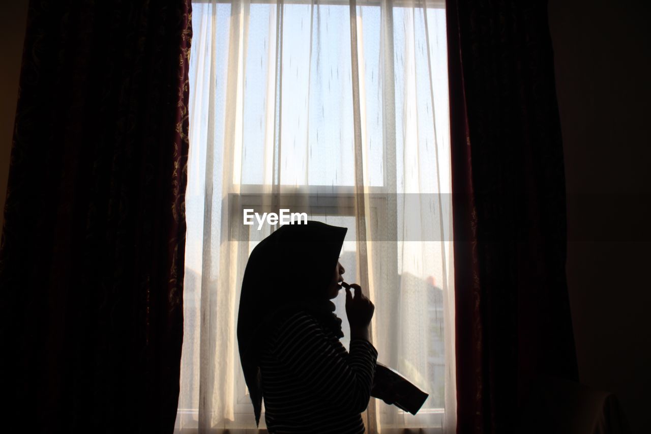 Side view of woman standing by window at home