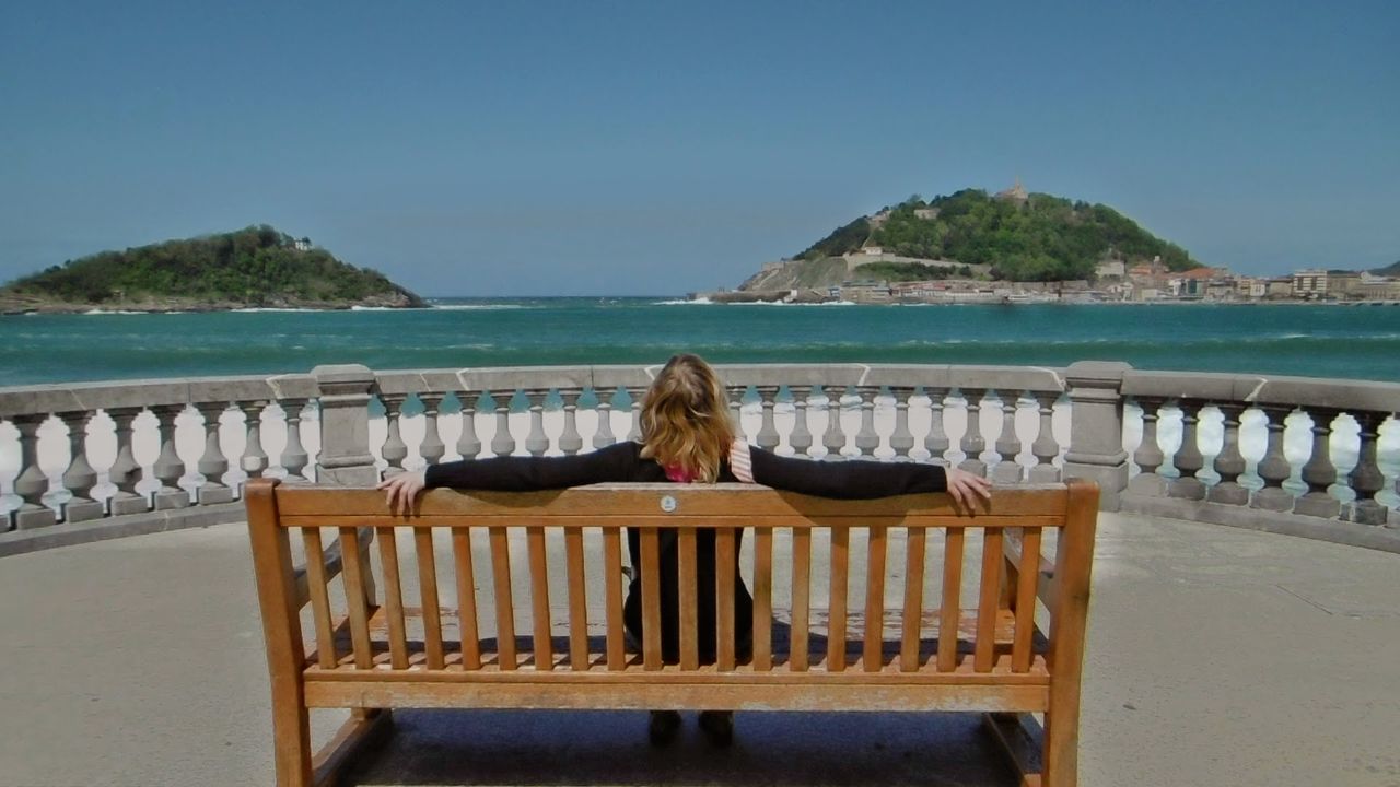 Rear view of woman relaxing on beach against clear blue sky