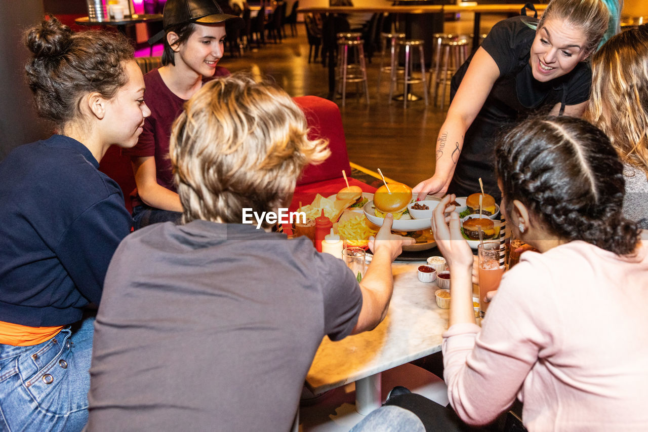 Smiling waitress serving food to multi-ethnic teenagers sitting at restaurant