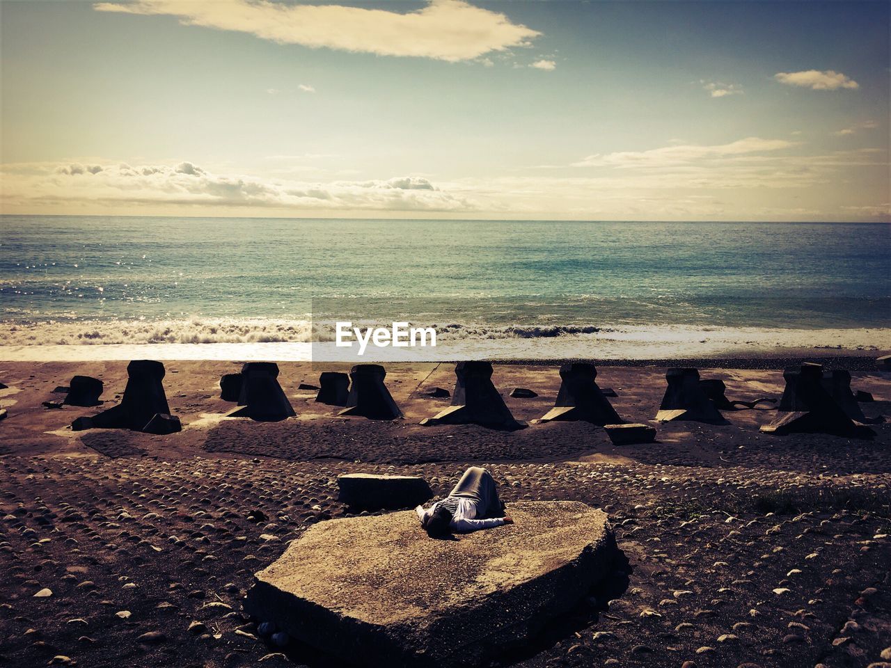 Woman sleeping on rock at beach against cloudy sky