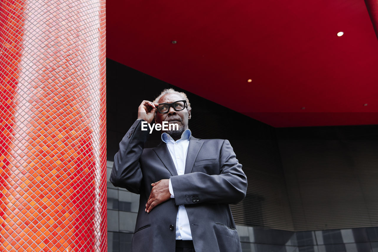 Senior male professional adjusting eyeglasses while standing by red column