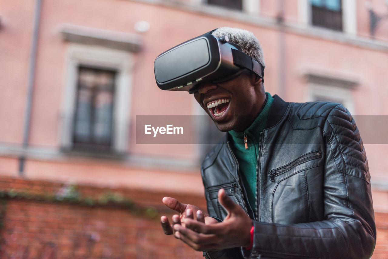 Excited african american male in vr goggles experiencing virtual reality and laughing in city street