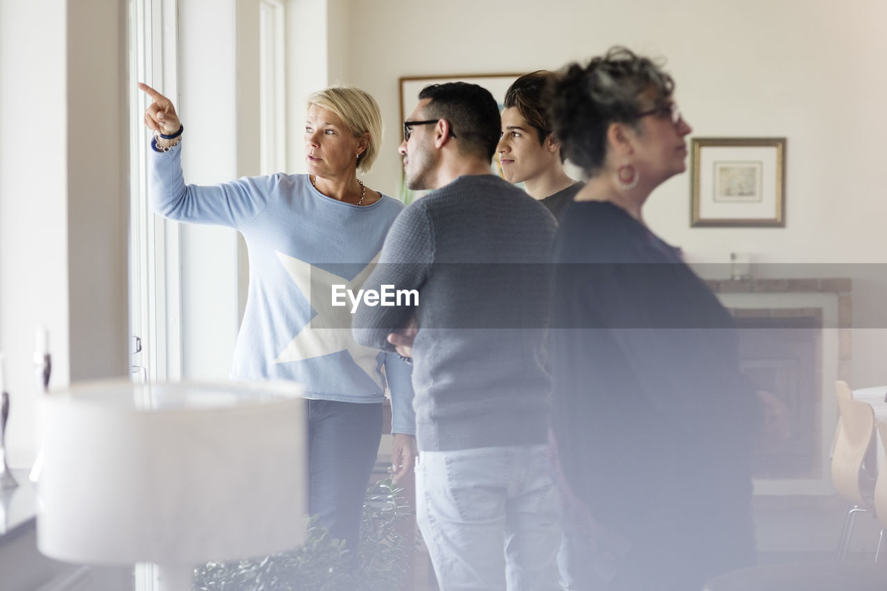 Mature woman pointing and showing to friends through window while standing in living room