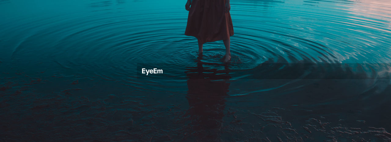 Low section of woman standing in sea during sunset