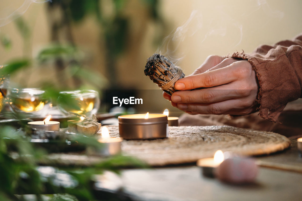 Woman hands burning white sage, before ritual on the table with candles and green plants. smoke of