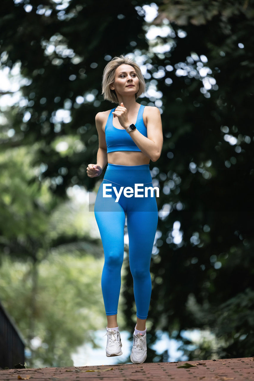 A beautiful slim blonde is running through a green park in a big city wearing blue leggings.
