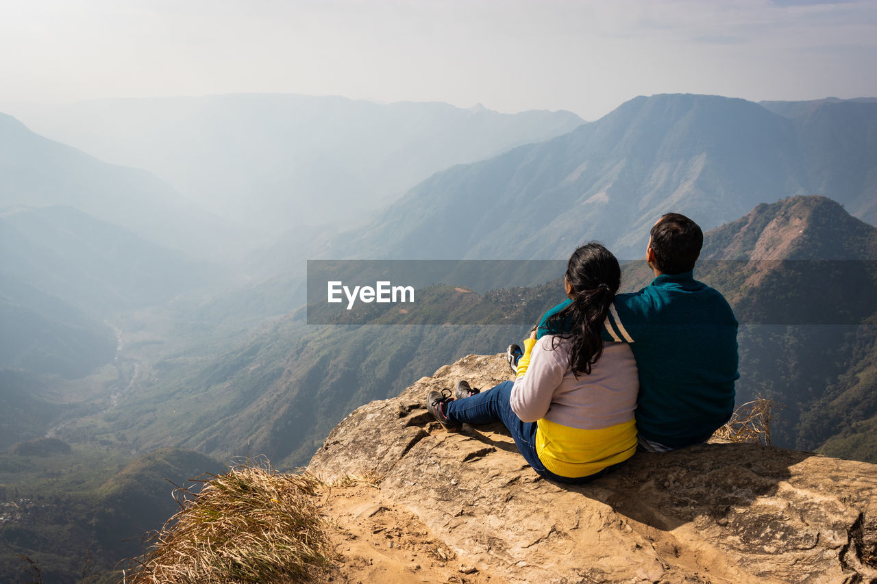 Rear view of couple sitting on cliff against mountains