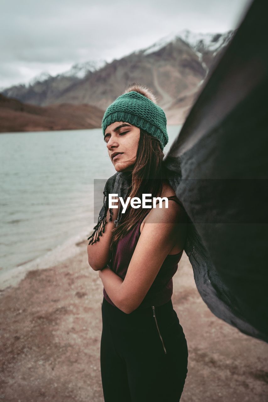 YOUNG WOMAN WEARING HAT STANDING BY MOUNTAIN