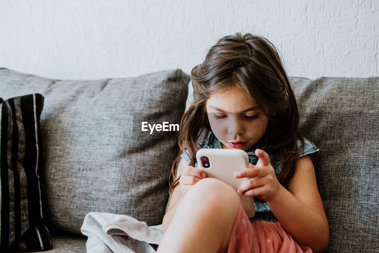 Adorable ethnic little girl in casual clothes and socks relaxing on comfortable sofa and playing video game with smartphone