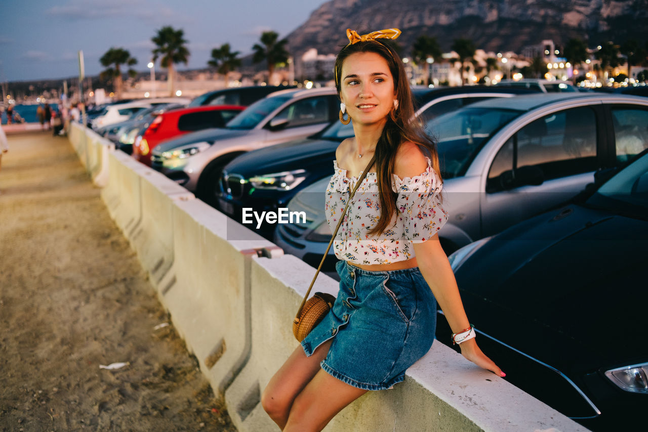 FULL LENGTH OF WOMAN SMILING WHILE SITTING ON CAR