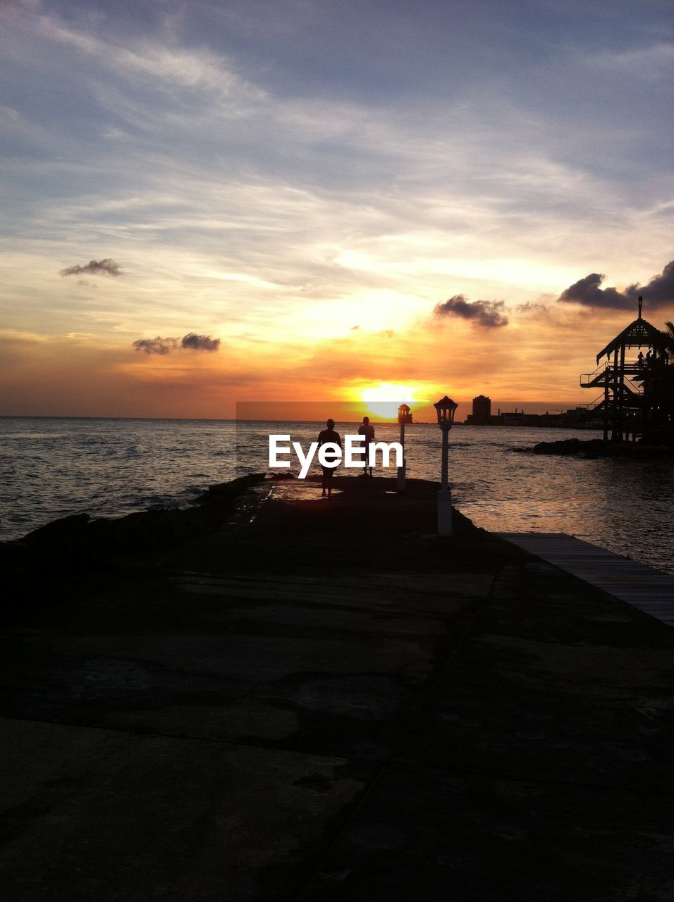 Silhouette man and woman standing on jetty in sea against sunset sky