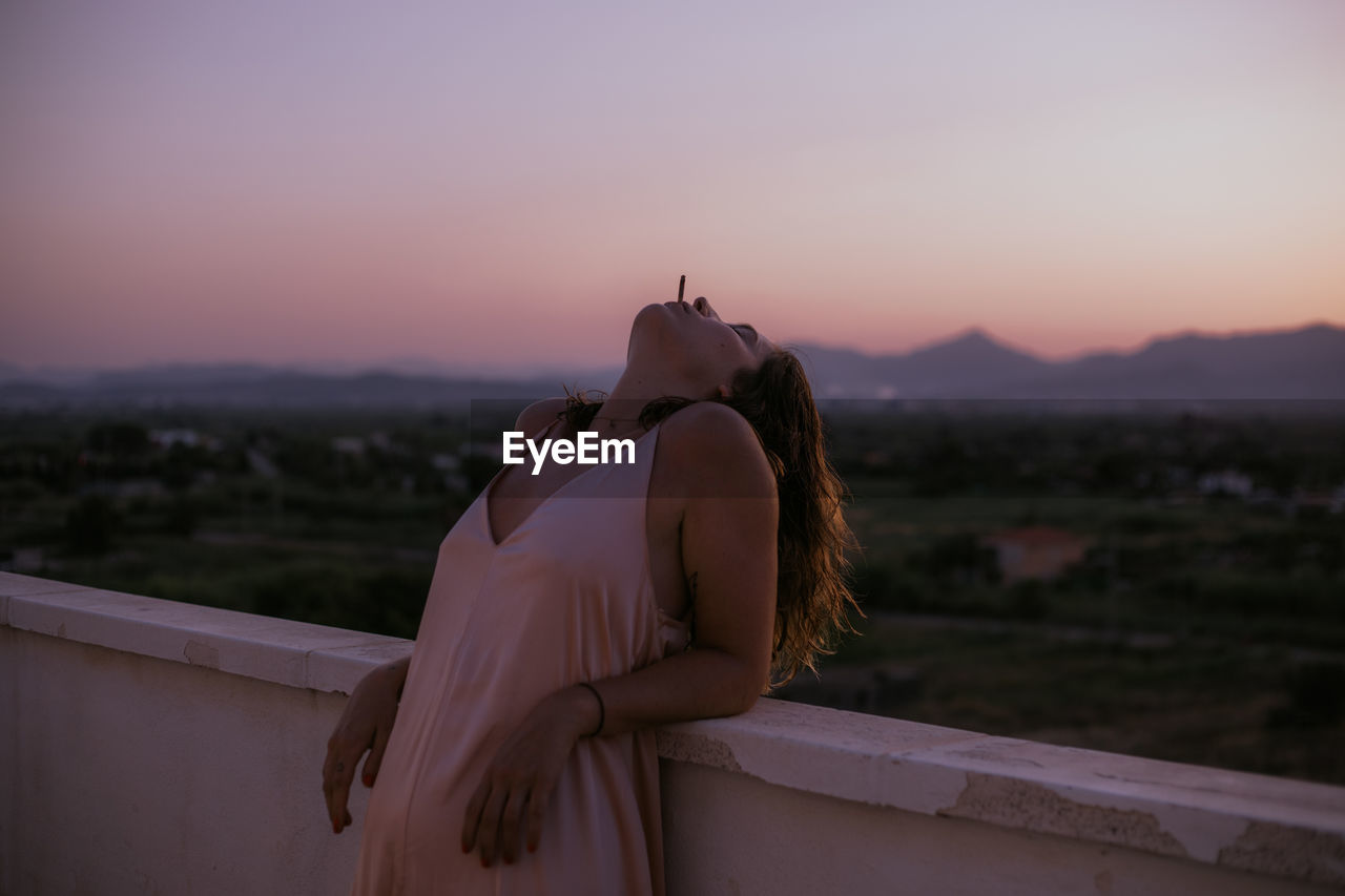 Side view of peaceful lady lying with closed eyes on fence of balcony and smoking cigarette with sunset on blurred background