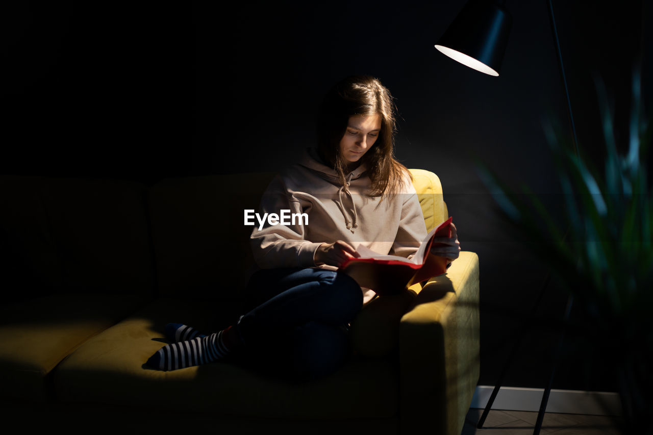 Full length of young woman reading book sitting on sofa