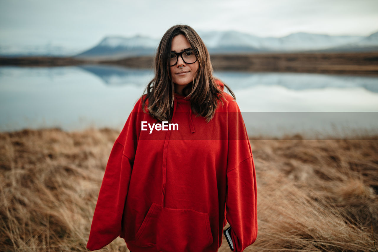 Young cheerful tourist in eyeglasses with piercing and windy hair near water in sunny day on blurred background