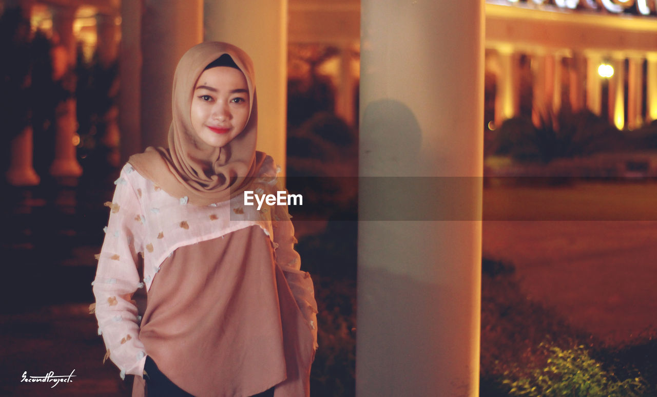 PORTRAIT OF SMILING YOUNG WOMAN STANDING OUTDOORS