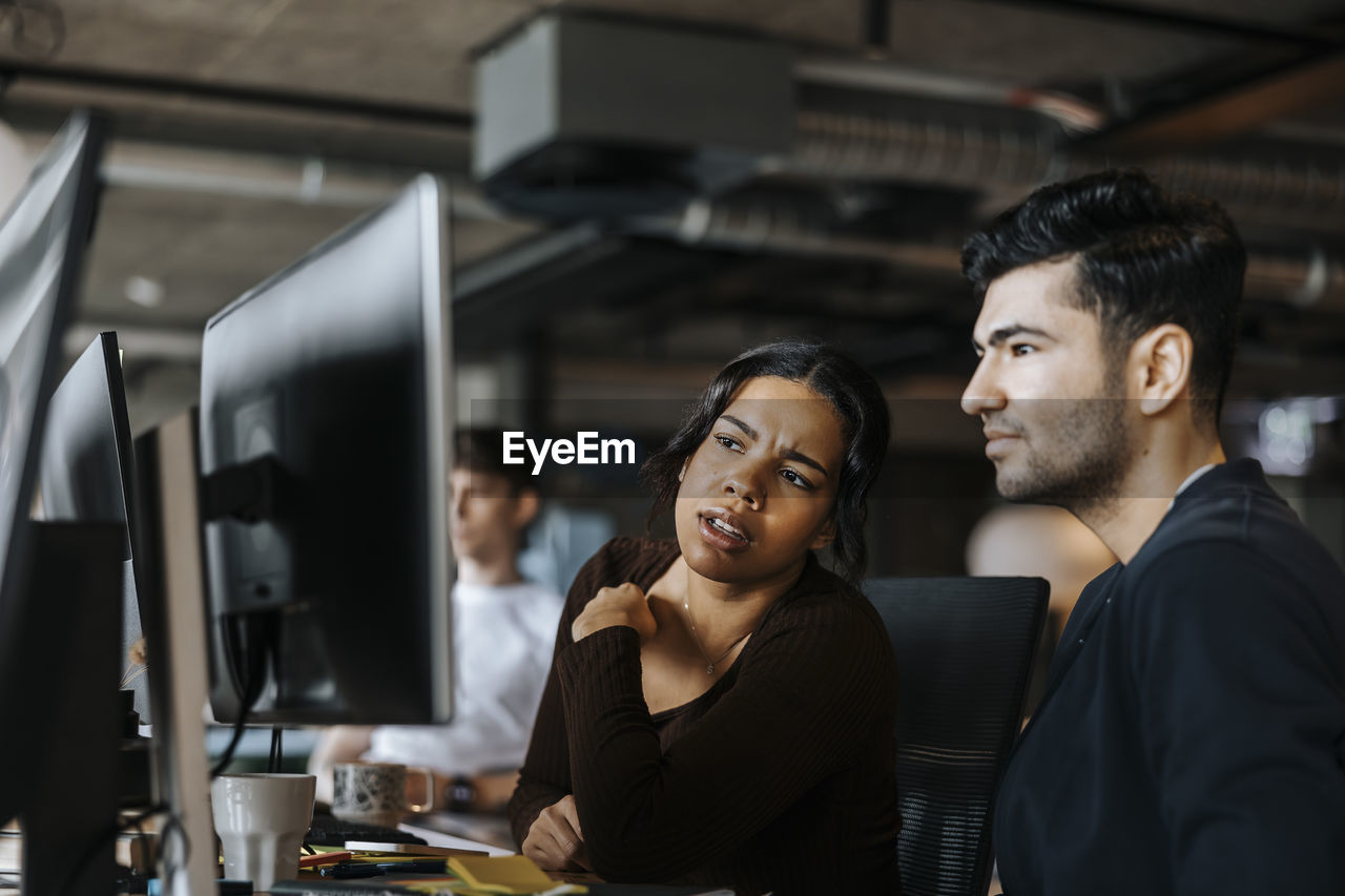 Young female programmer looking at monitor screen while sitting next to male colleague at office