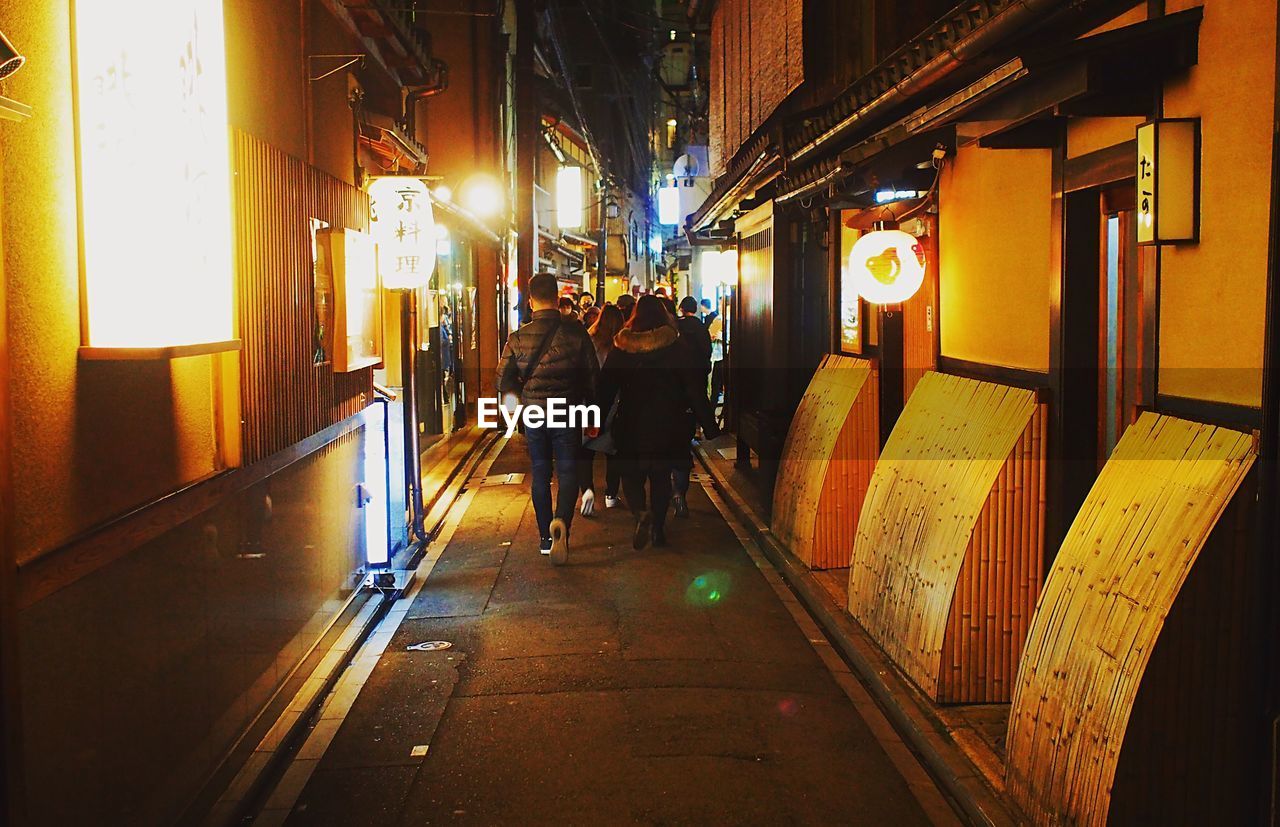 REAR VIEW OF PEOPLE WALKING ON ILLUMINATED CORRIDOR