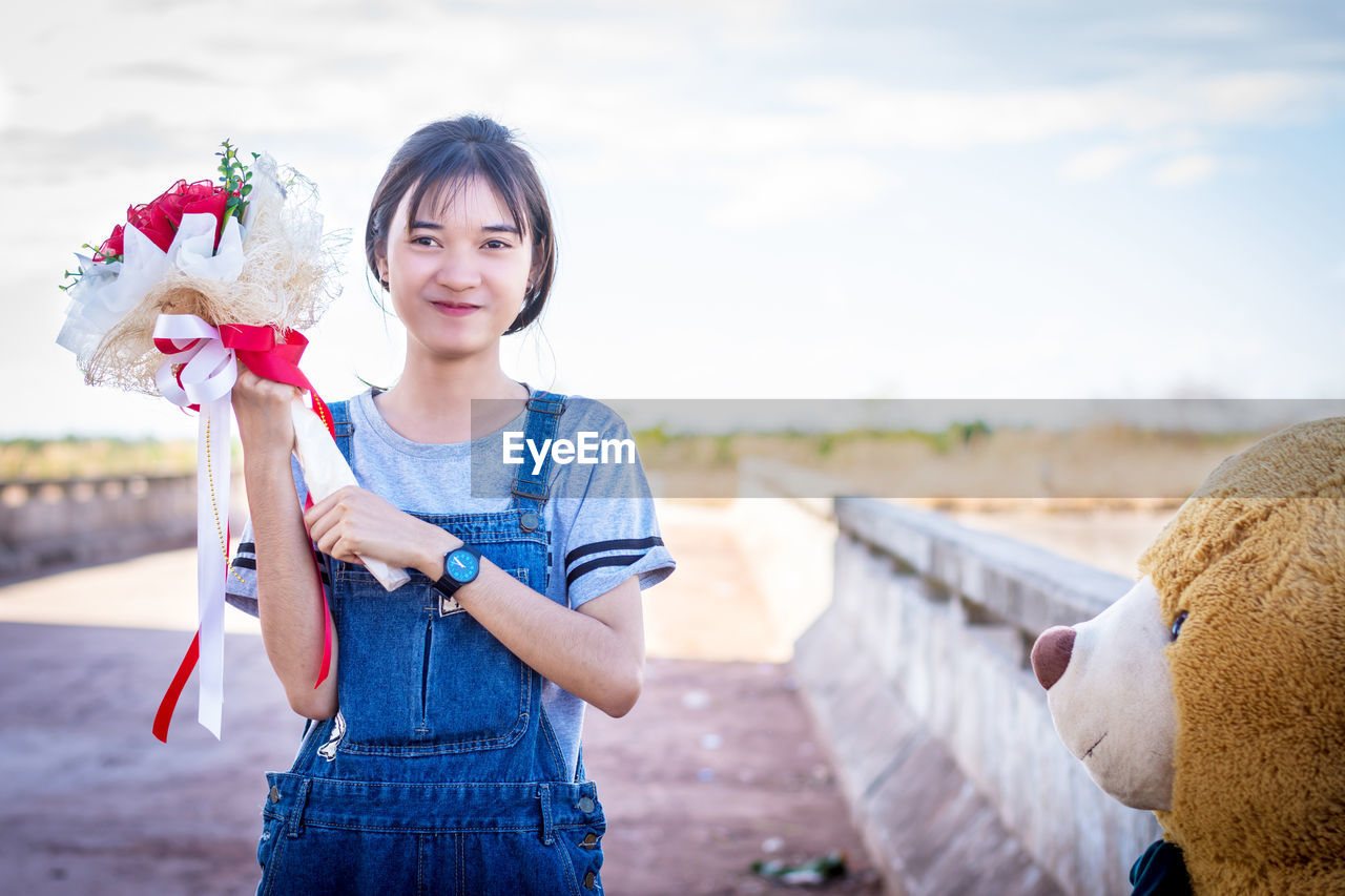 Portrait of young woman holding bouquet while standing against sky