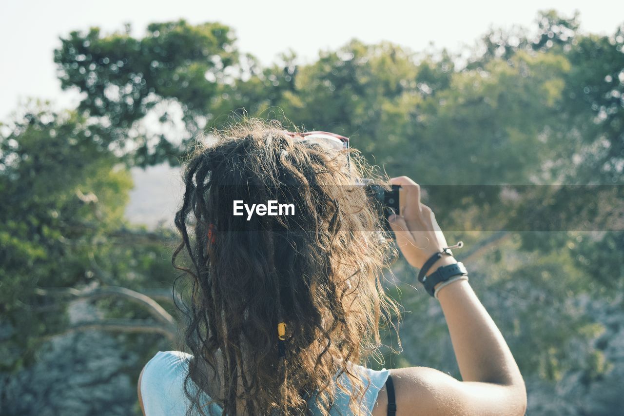 Rear view of woman photographing through mobile phone against trees