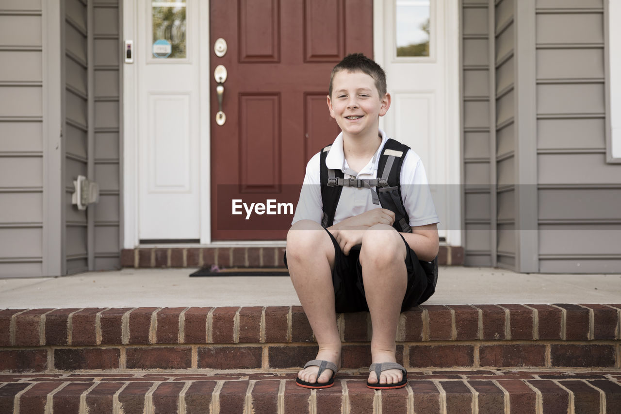 Tween boy with braces sits on brick front step, first day of school