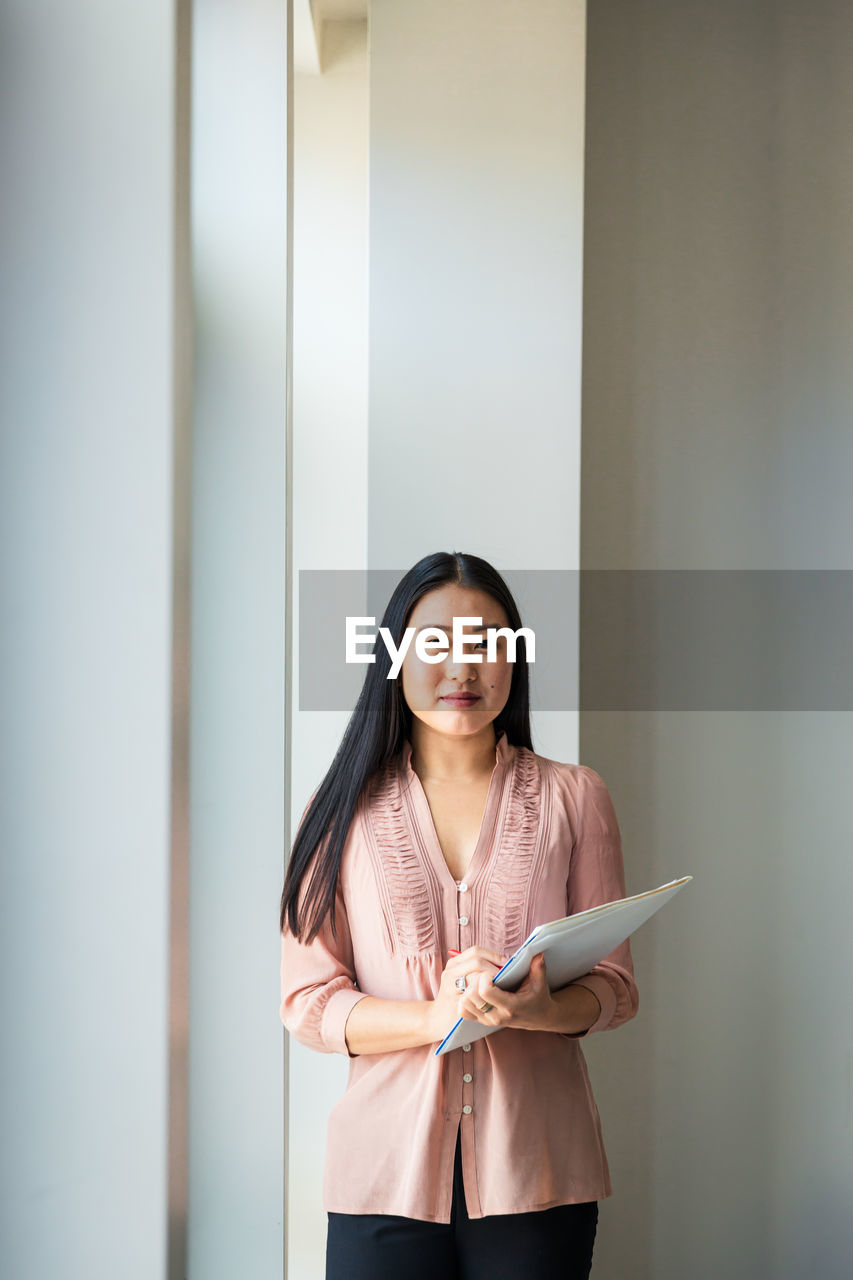 Portrait of confident businesswoman with file standing against wall in office