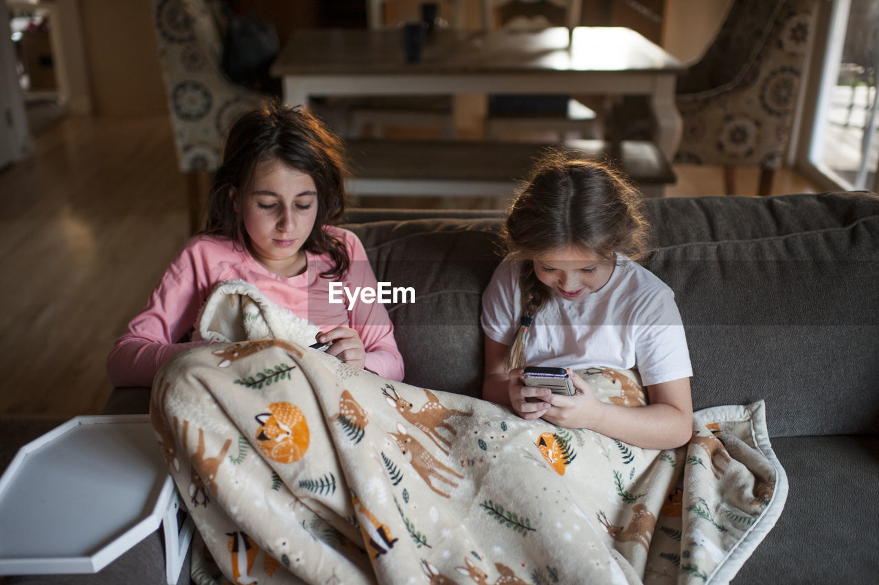 Friends sit on couch under blanket looking at their phones