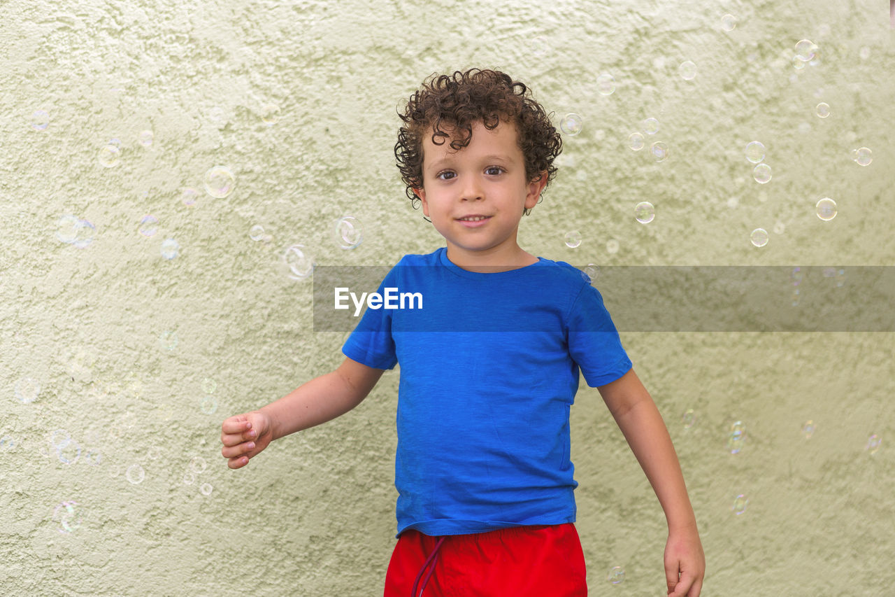 Cute boy with curly hair and caucasian appearance looking at the camera surrounded by soap bubbles