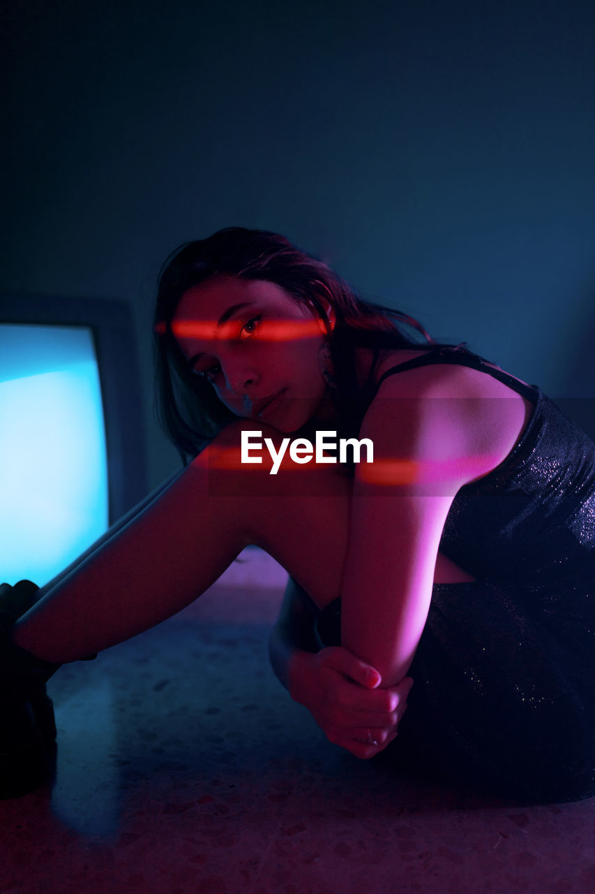 Side view of female model in black dress sitting looking at camera on floor near glowing old television in dark studio