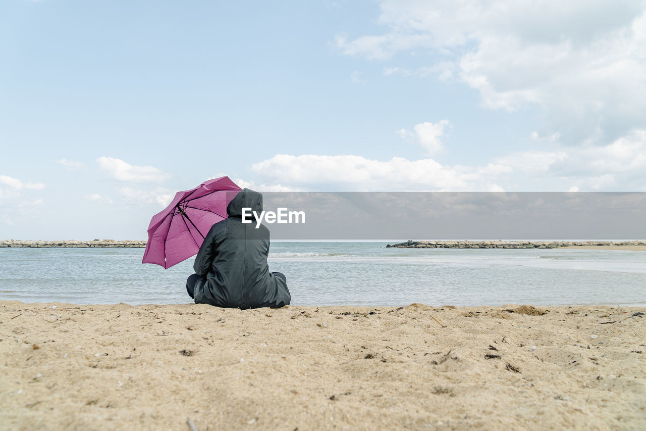 sky, beach, water, land, sea, shore, nature, one person, cloud, sand, ocean, coast, body of water, vacation, holiday, trip, adult, rear view, full length, beauty in nature, horizon, day, scenics - nature, leisure activity, horizon over water, relaxation, tranquility, sitting, women, solitude, outdoors, lifestyles, tranquil scene, copy space, environment, loneliness, travel, casual clothing, umbrella, summer, protection, clothing, travel destinations, men, person