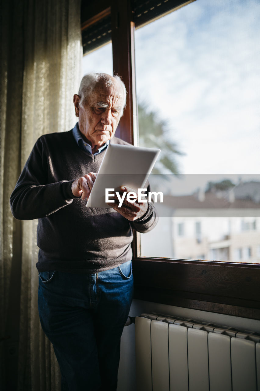 Senior man with tablet standing in front of window at home