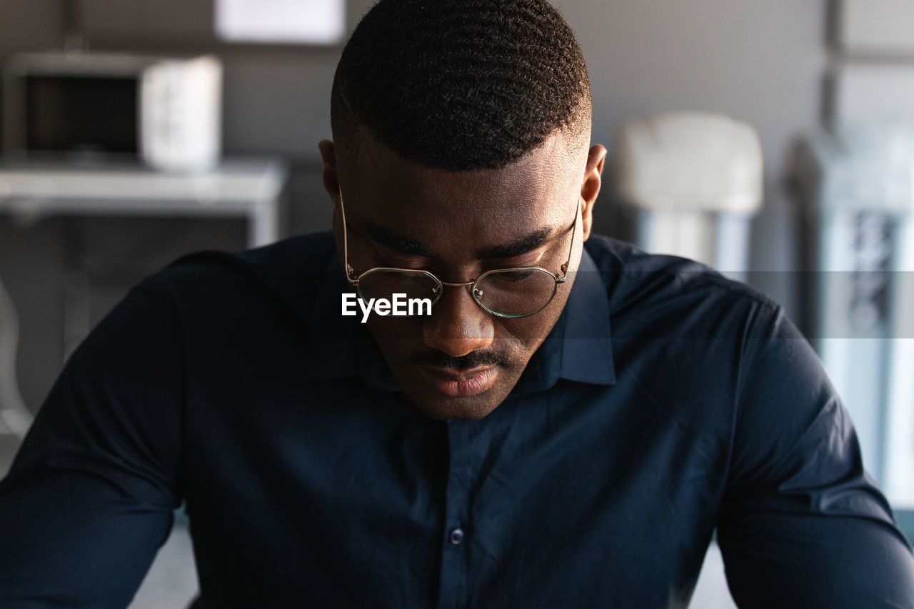 African american adult male in formal clothes and eyeglasses sitting at table and browsing on tablet near netbook and documents with takeaway drink in bright office