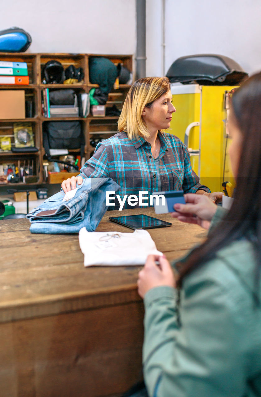 Female client holding credit card to pay purchase while woman shop assistant checking price of jeans