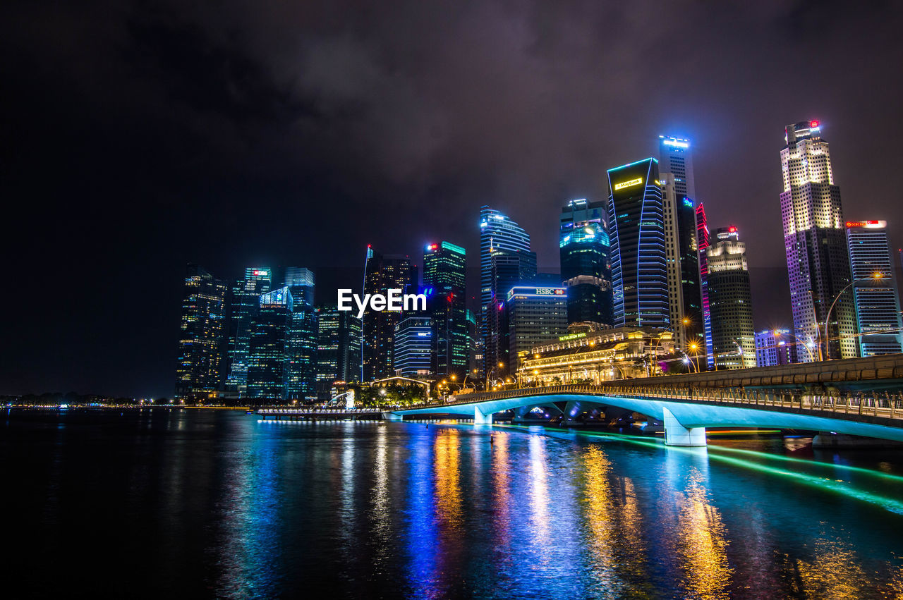 Illuminated buildings by river against sky at night