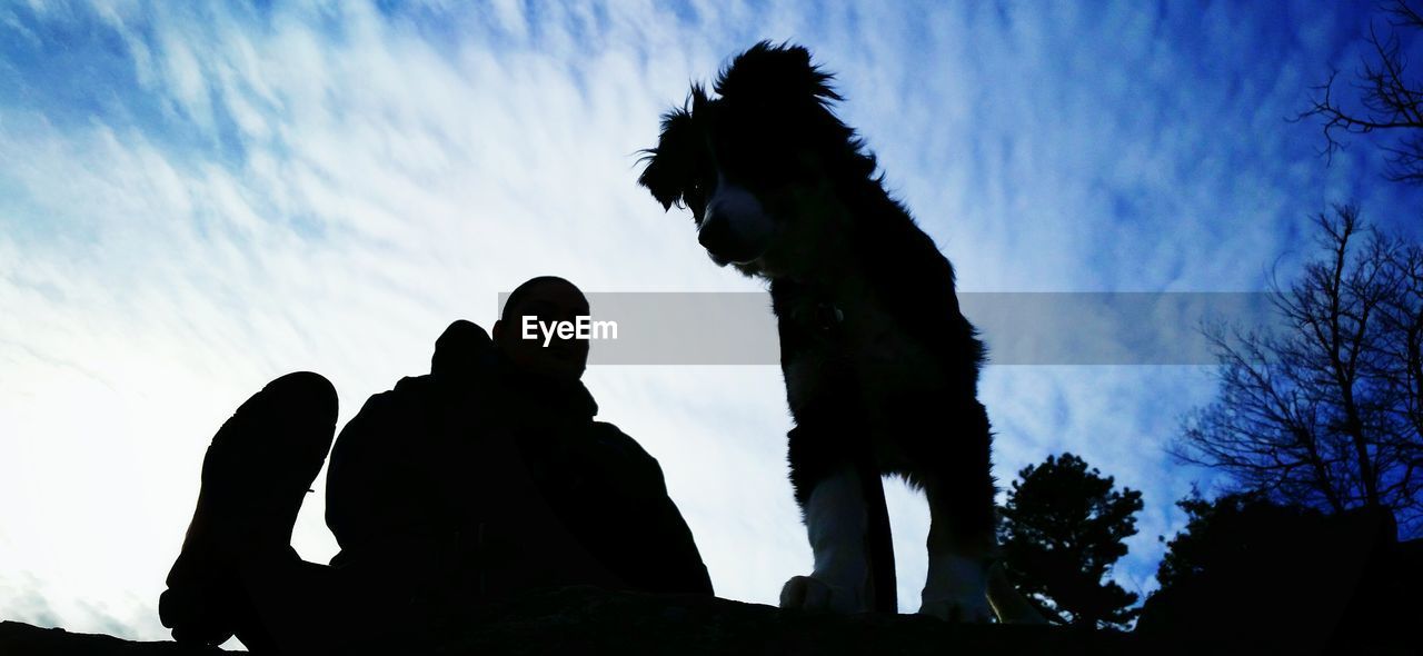 Low angle view of silhouette man and dog sitting against cloudy sky