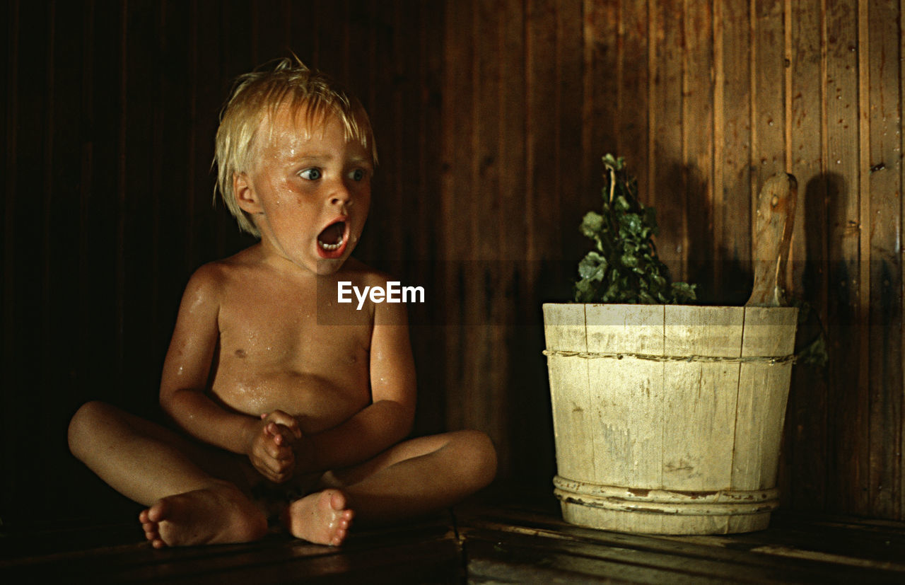 Scared boy looking away while sitting by potted plant against wall