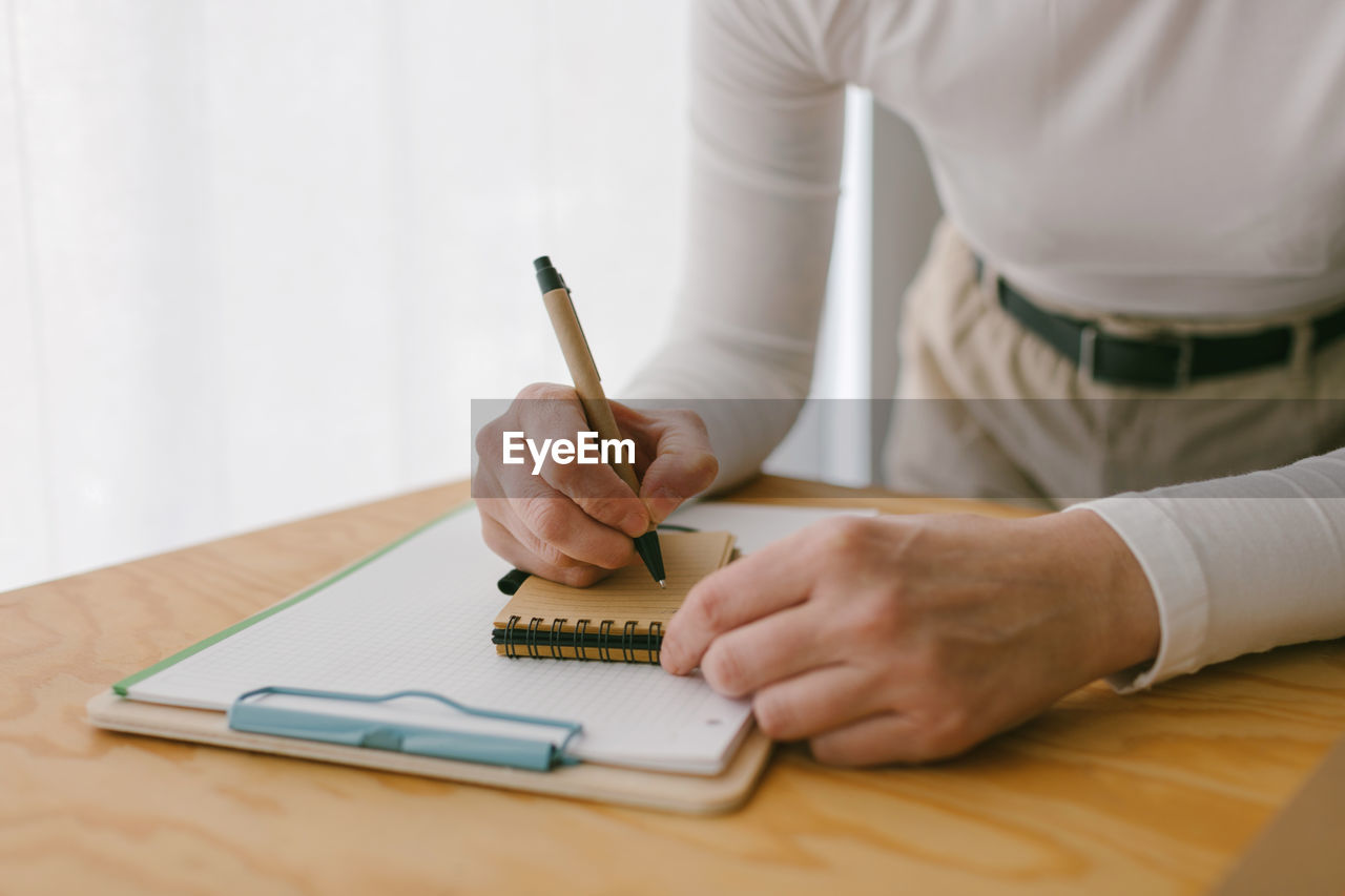 Faceless casual businesswoman bending on wooden table and writing in notepad with pen