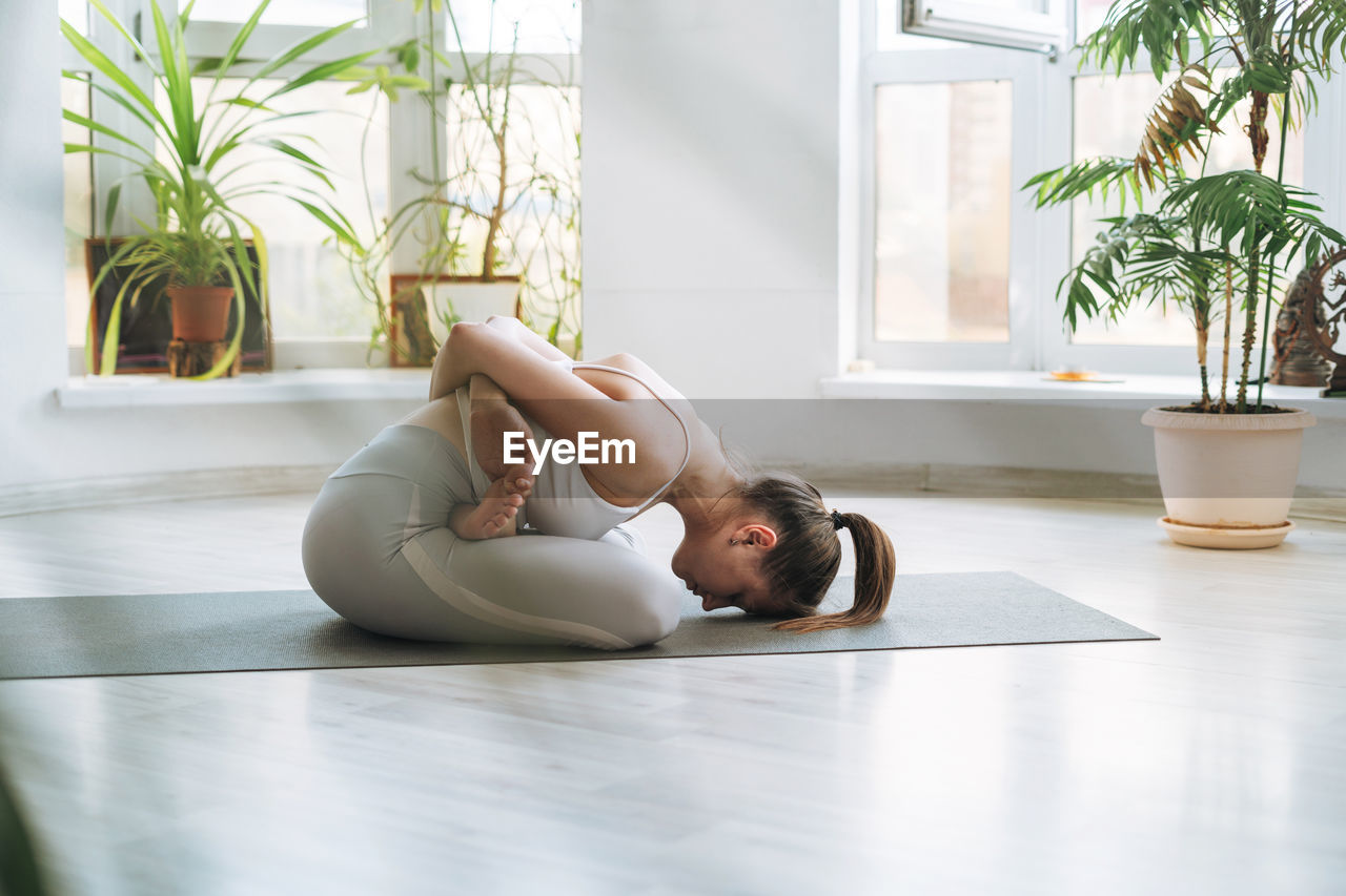 Young fit woman practice yoga doing asana in light yoga studio with green house plant