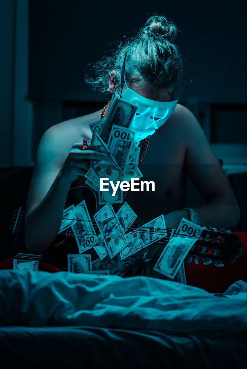 Young man wearing mask while sitting with money and guitar on bed