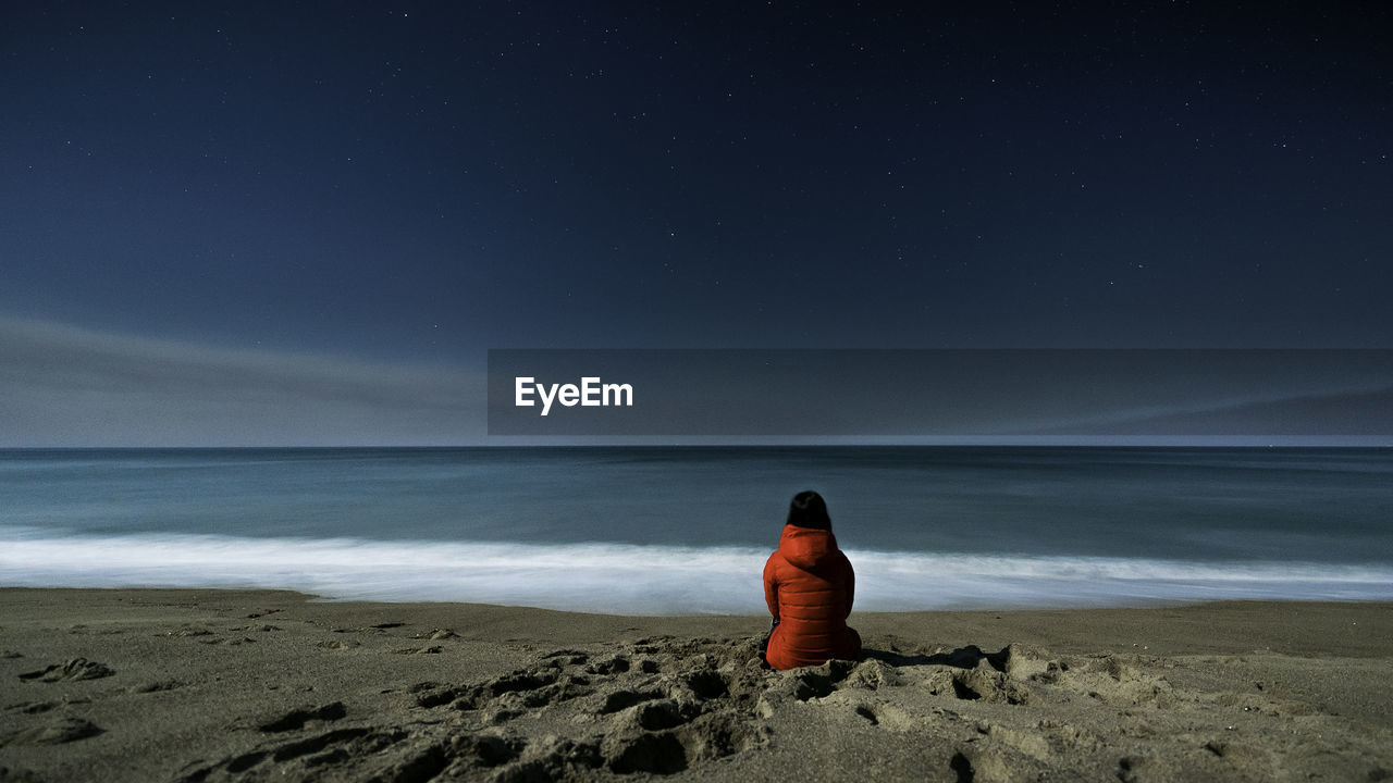 Rear view of woman sitting at beach against sky at night